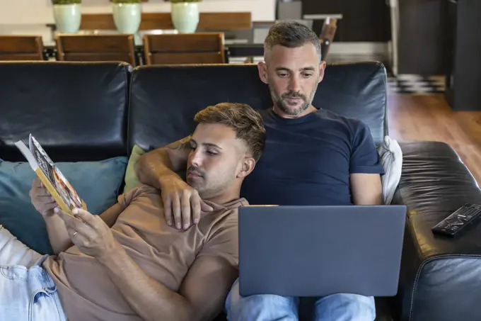 Gay male couple.  One is working and the other one is reading a book