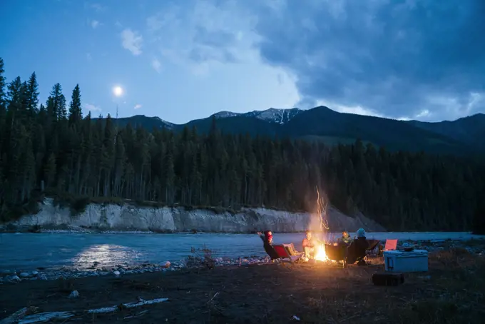 A group of adults sit around campfire beside wilderness river