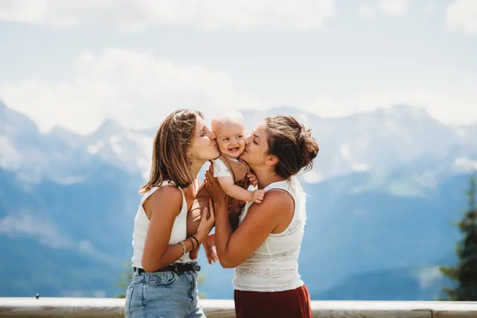 Two moms kissing smiley baby on cheek in summer in mountains