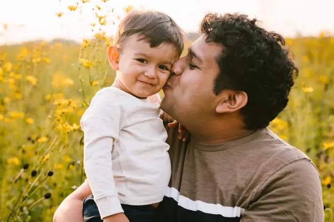Toddler son smiles while Latino Dad kisses cheek outside in field