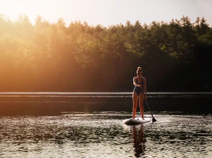 Woman in swim suit using paddleboard on lake in sunshine.