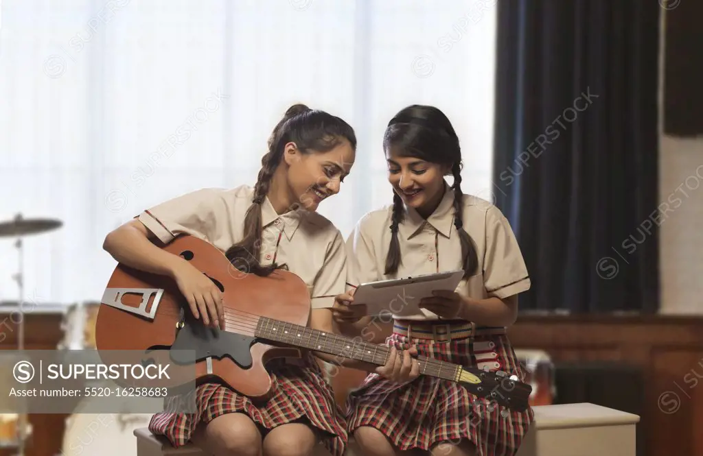 Two happy teenage school girls learning to play guitar looking at musical notes in the music room at school
