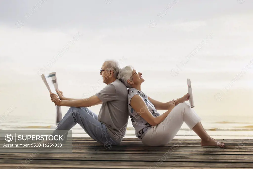 Senior couple sitting back to back on wooden dock near lake and reading newspaper