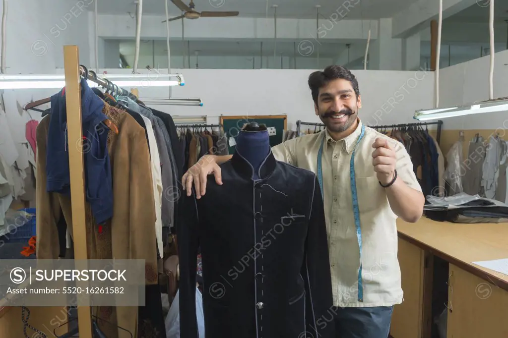 Tailor standing beside a mannequin wearing a semi-stitched coat in his workshop and showing a gesture of holding a product in hand.