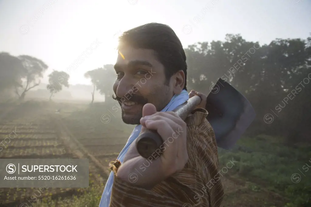 Side view of a smiling farmer walking in his agriculture field carrying a spade on his shoulders with sun in the background.