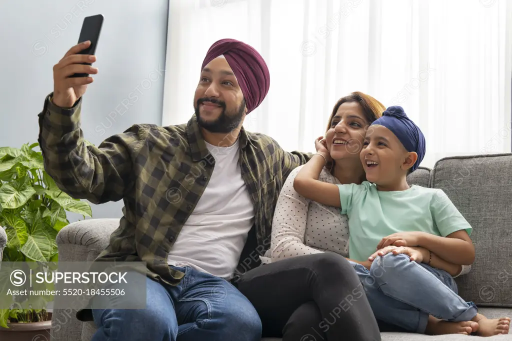 A SIKH SON HUGS MOTHER WHILE FATHER CLICKS A SELFIE OF EVERYONE POSING TOGETHER ON A SOFA