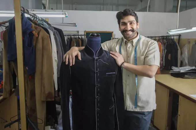 Tailor standing beside a mannequin wearing a semi-stitched coat in his workshop.