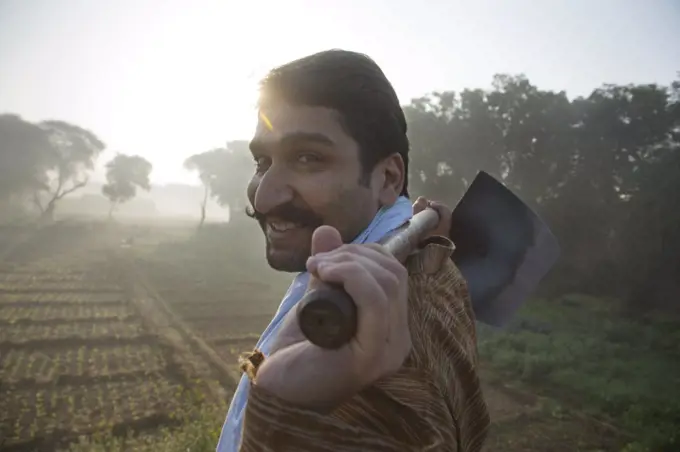 Side view of a smiling farmer walking in his agriculture field carrying a spade on his shoulders with sun in the background.