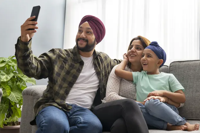 A SIKH SON HUGS MOTHER WHILE FATHER CLICKS A SELFIE OF EVERYONE POSING TOGETHER ON A SOFA