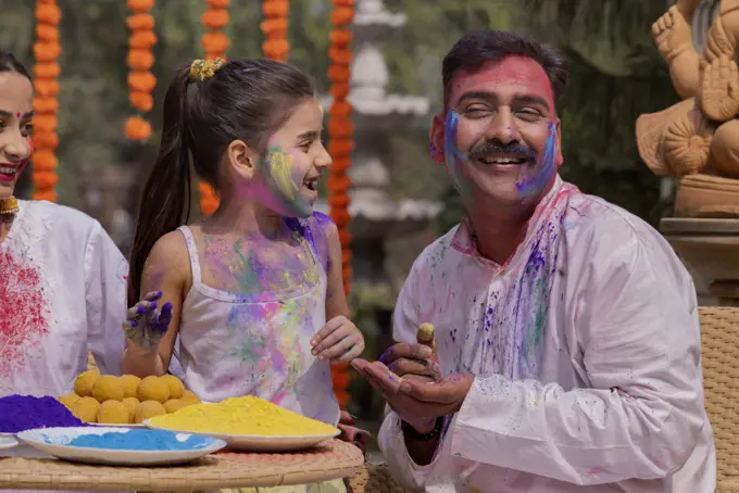 Granddaughter applying colour on grandfather's face and mother sitting beside on the occasion of Holi