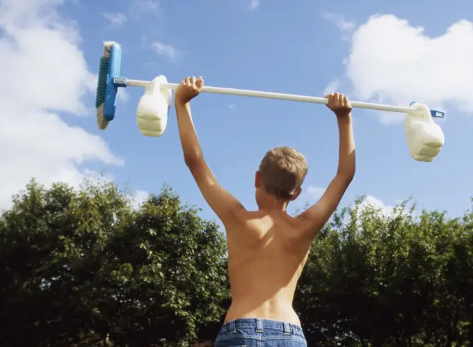 Back view of young boy lifting full 2 litre bottles of milk on broom