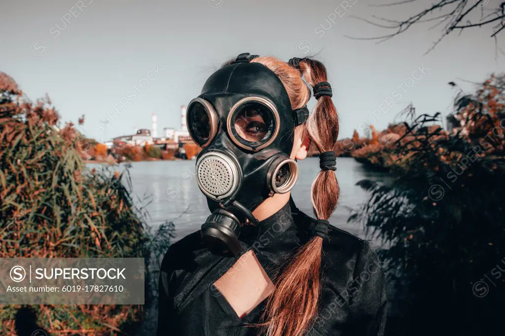 young girl in a gas mask in a red dress with a book in her hands against the background of smoking factory chimneys