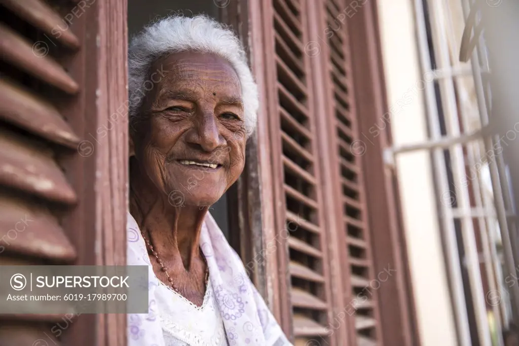 An old Cuban woman looks out of a window with shutters and smiles.