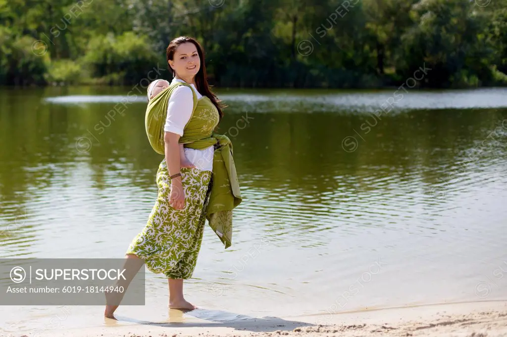 Mother with wrapping infant baby in green sling back at nature