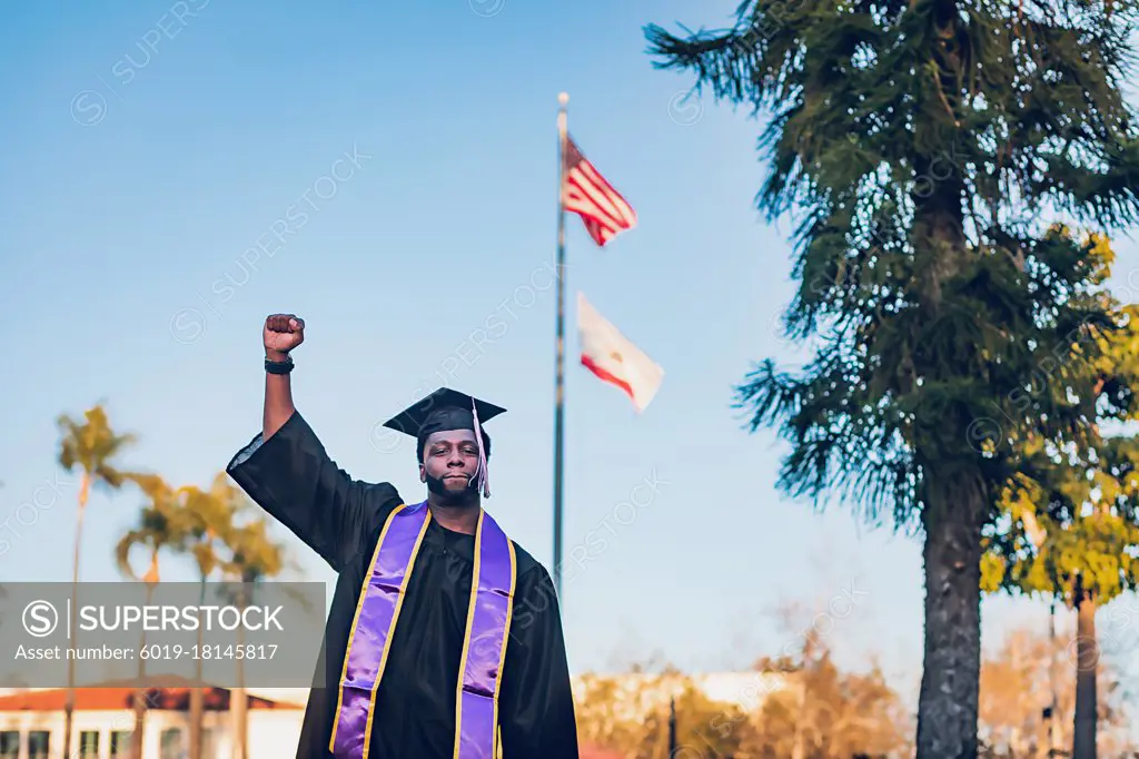 Young man wearing a graduation gown/cap in front of an american flag.