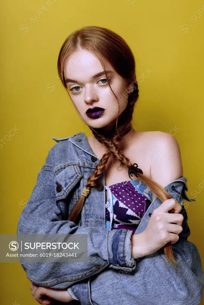 A young beautiful red haired girl posing in jeans jacket with purple lips on yellow background.