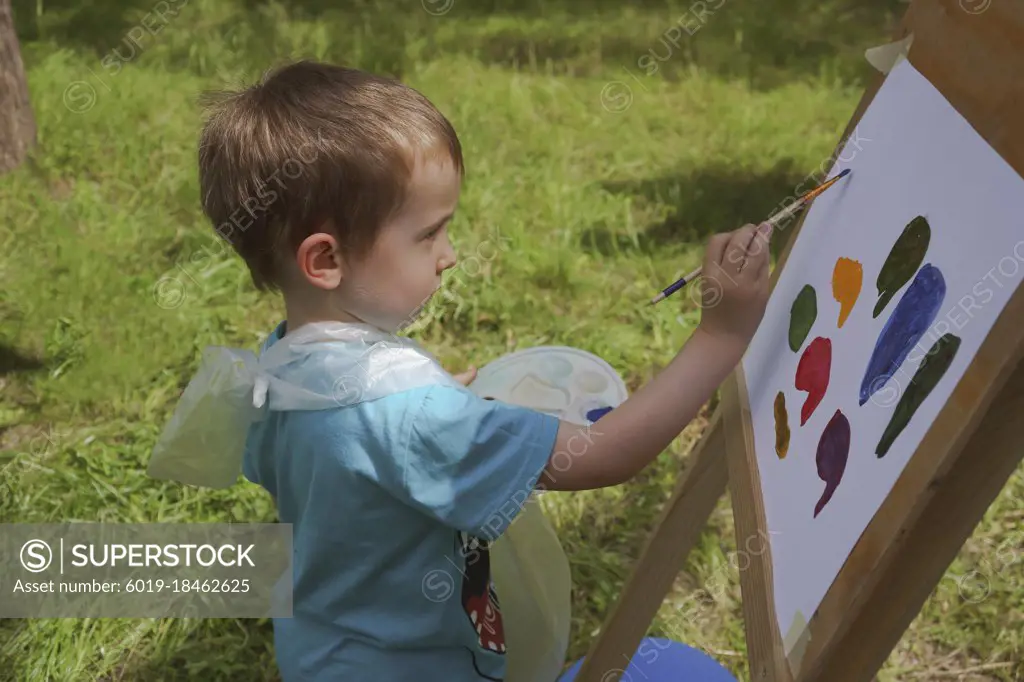 boy 3-4 years old draws on an easel with paints diligently outdoors