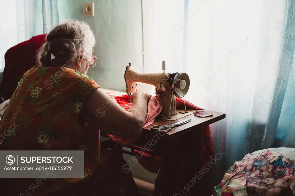 Old gray-haired woman with curly hair sews on retro sewing machine