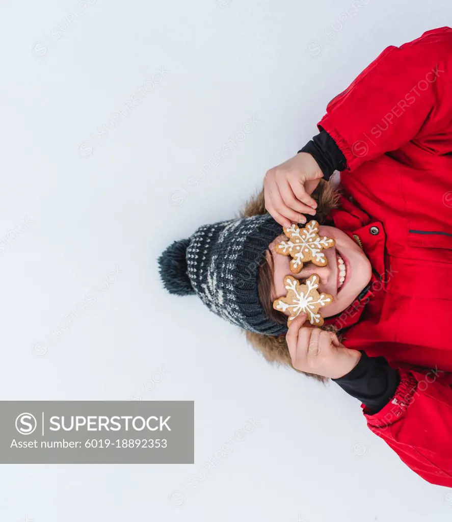 Overhead of boy with snowflake cookies over eyes laying in the snow.