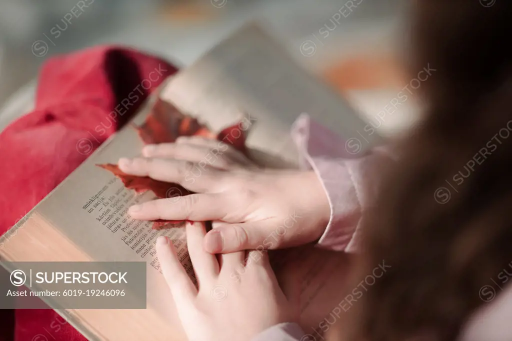 The girl is reading a book and holding a red maple leaf in her hand.