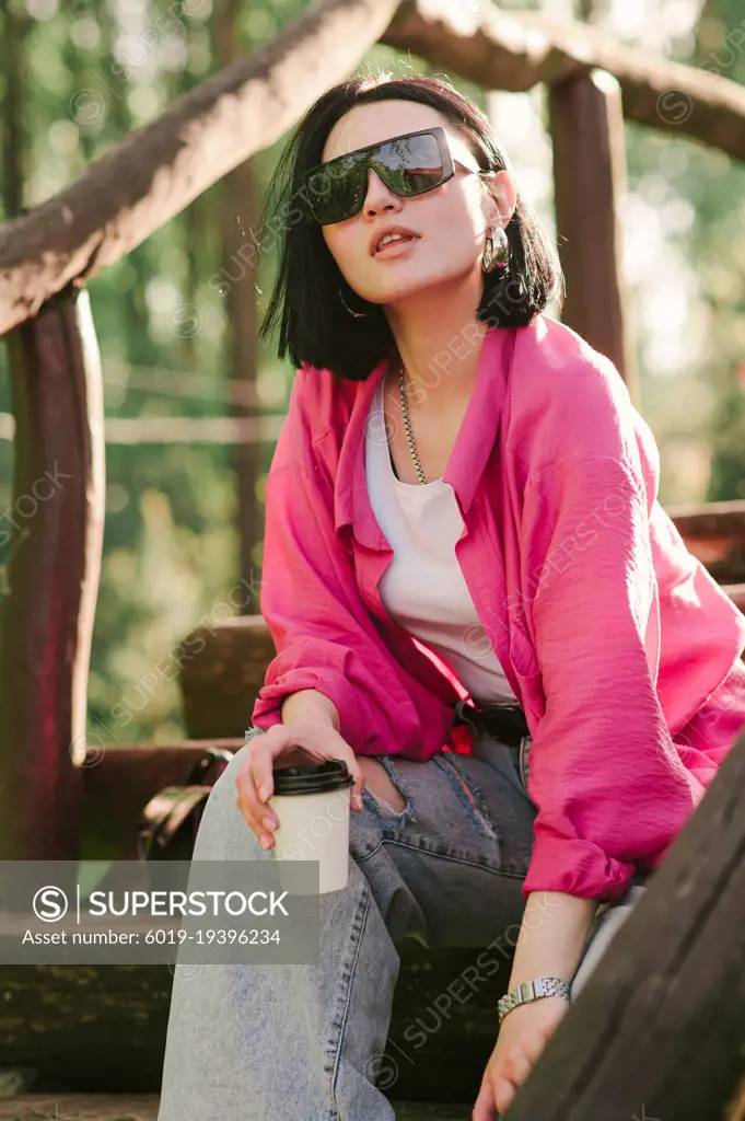 Stylish woman in sunglasses holding cup of coffee outdoors in the park