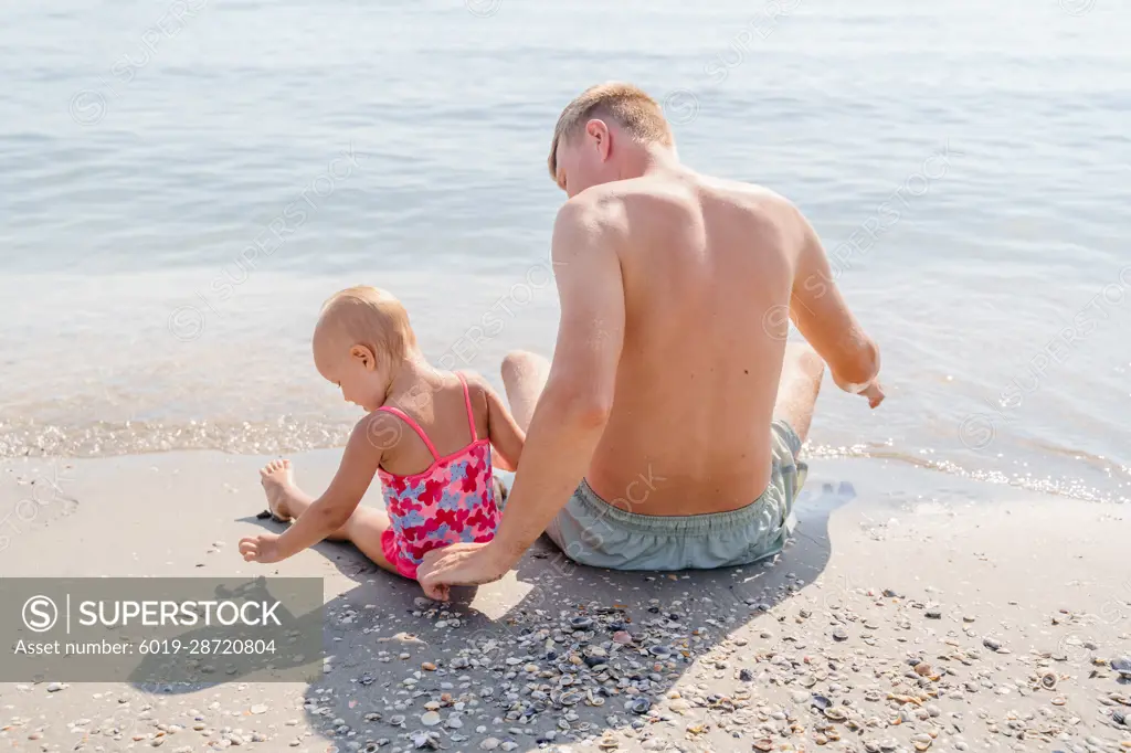 Girl baby toddler with dad sit on the beach by the sea, back view