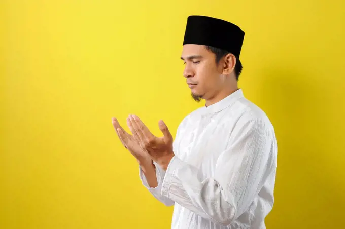 Young Asian Muslim man praying raising his hand with moslem clothes, isolated on yellow background