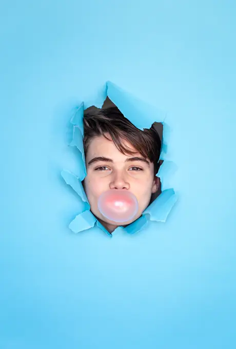Boy blowing bubble with head through hole in blue paper background.
