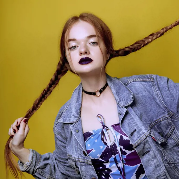 A young beautiful red haired girl posing in jeans jacket with purple lips on yellow background.