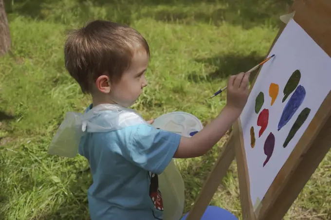 boy 3-4 years old draws on an easel with paints diligently outdoors