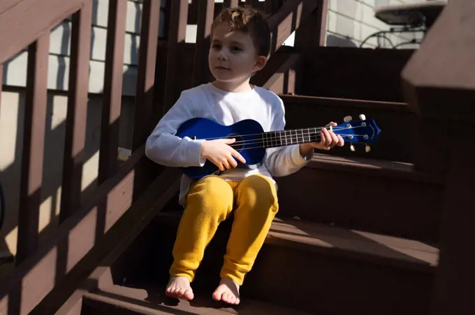 Little boy playing a blue ukulele on the back steps