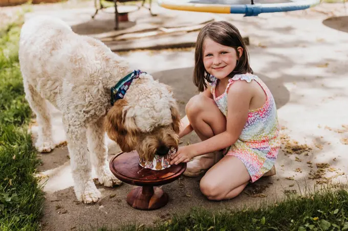 Young girl holding cake while dog eats it outside in backyard