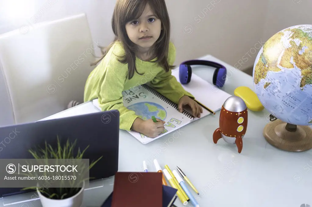 Smiling child girl draws planet earth with wax colors on school notebook for Earth day - Little activist girl writes the message Save the Planet - Protection of environment and global warming concept
