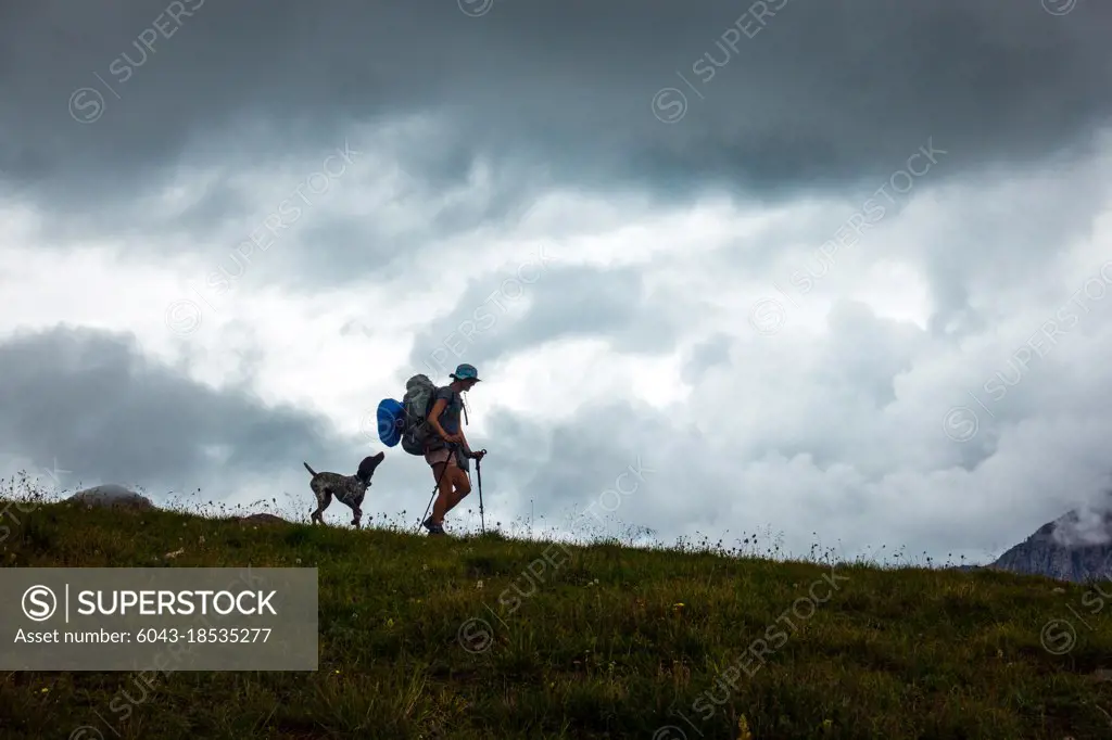 Woman hiking along ridge with poles as her dog follows closely behind at sunset in Alpine Colorado