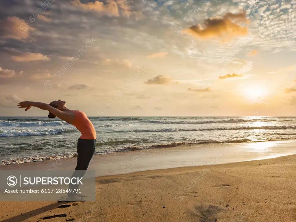 Young sporty fit woman doing yoga Sun salutation Surya Namaskar pose Hasta Uttanasana on tropical beach on sunset
