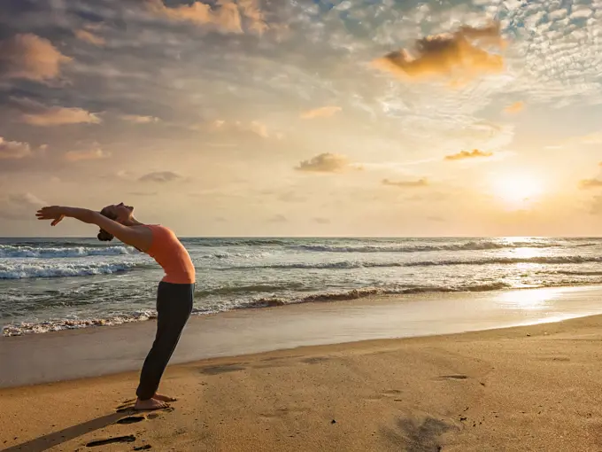 Young sporty fit woman doing yoga Sun salutation Surya Namaskar pose Hasta Uttanasana on tropical beach on sunset