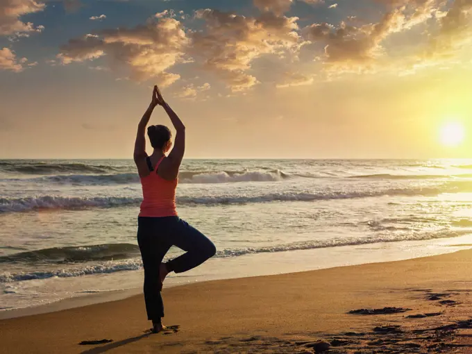 Yoga outdoors - sporty fit woman doing Hatha yoga asana Vrikshasana tre pose on tropical beach on sunset