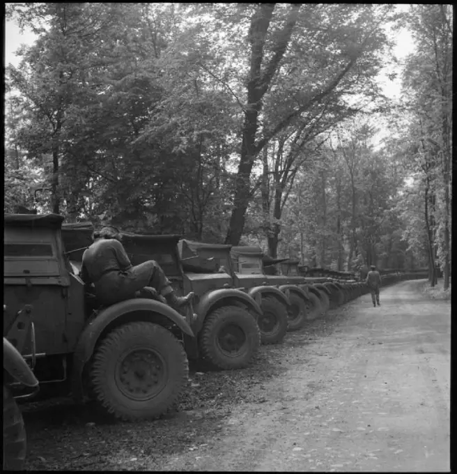 Invasion Build-up- Preparations For the D-day Landings, UK, 1944 A line of army trucks awaits collection along a tree-lined lane or path, somewhere in Britain. They will soon be collected by various units and transported to the Second Front. In the foreground, an ATS mechanic can be seen sitting on the wheel arch of one of the vehicles, her back to the camera. Another member of the ATS can be seen walking down the lane into the distance.