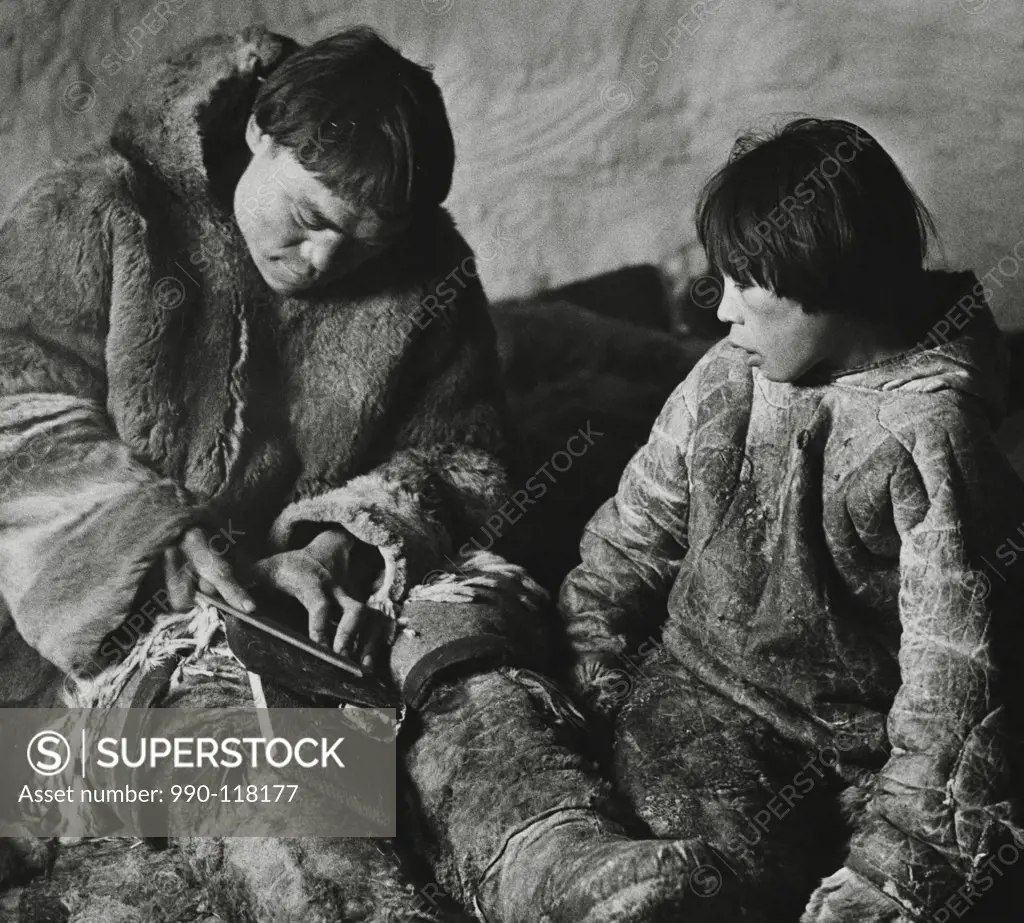 Mid adult Eskimo man sharpening an axe with his son sitting beside him