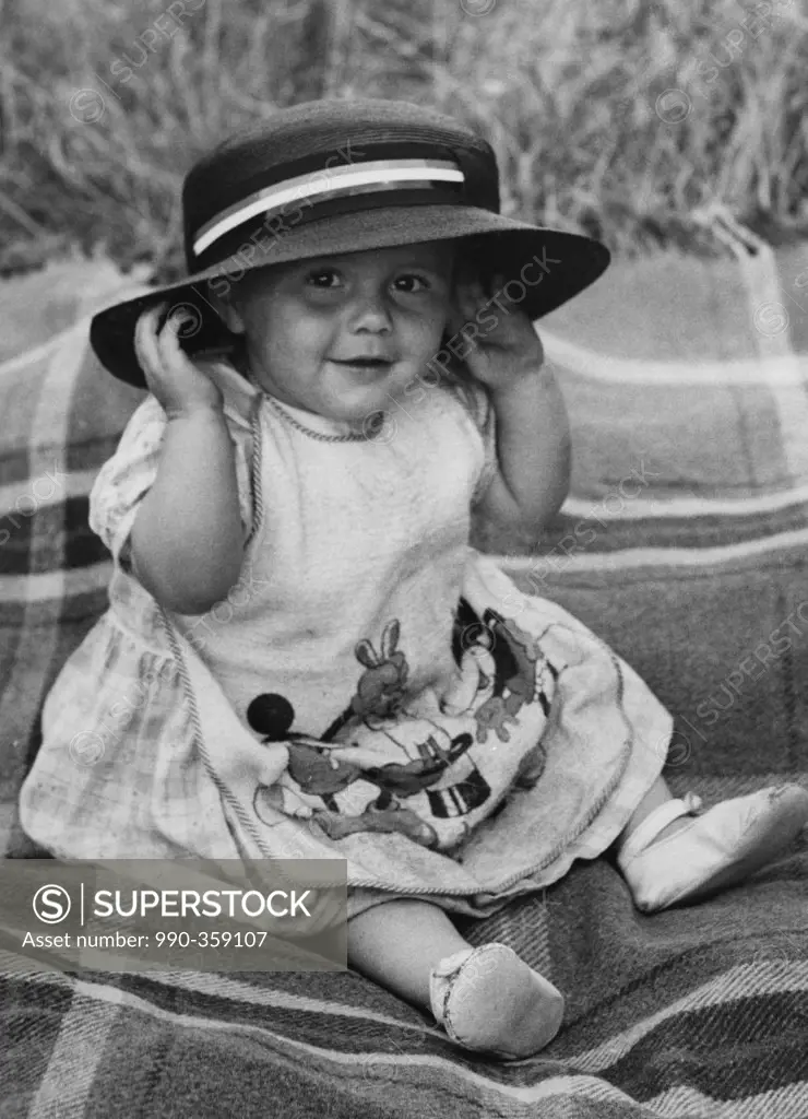 Baby girl sitting on blanket, smiling and wearing hat